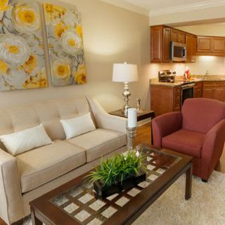 A cozy living room connected to a kitchen, featuring a comfortable sofa, red armchair, decorative artwork, and a glass-top coffee table with a lamp.