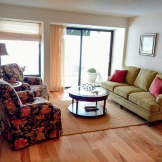 A cozy living room featuring a green sofa, floral armchairs, and a round coffee table, illuminated by natural light from the window.