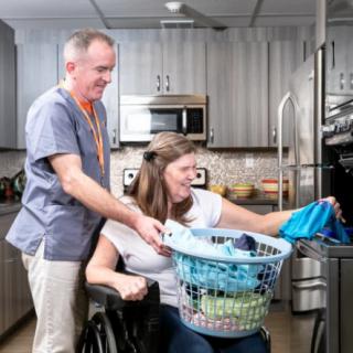 A caregiver assisting a person in a wheelchair with laundry in the kitchen.