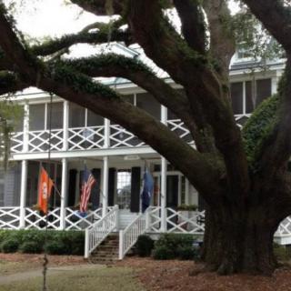 A large tree stands in front of a two-story house with a spacious porch.