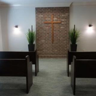 A simple chapel with wooden pews facing a brick wall adorned with a cross, flanked by plants and illuminated by wall lights.