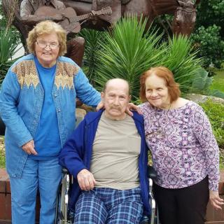Three elderly individuals, two standing and one seated in a wheelchair, enjoy time together outdoors.