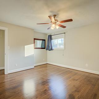 An empty room with wooden flooring and a ceiling fan, featuring large windows that let in natural light.