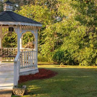A charming gazebo surrounded by a lush garden and trees in the warm sunlight.