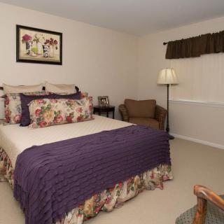 A cozy bedroom with floral-patterned bedding and a purple throw blanket, featuring a lamp and a comfortable chair in the corner.