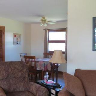 A cozy living room with a view of the adjoining dining area, featuring comfortable sofas, a table lamp, and a window bringing in natural light.