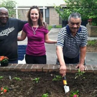Three people are enjoying a community gardening activity, smiling while planting flowers in an outdoor setting.