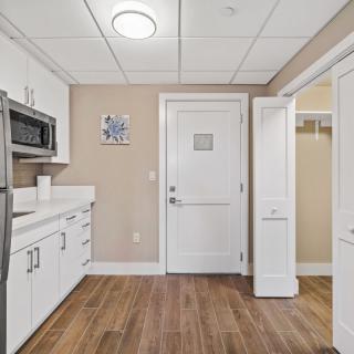 A modern kitchen area with white cabinets and a microwave, featuring wooden flooring and a door leading to another room.