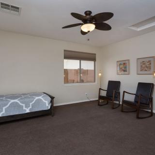 A minimalist bedroom featuring a single bed, two rocking chairs, a ceiling fan, and a window with framed wall art.