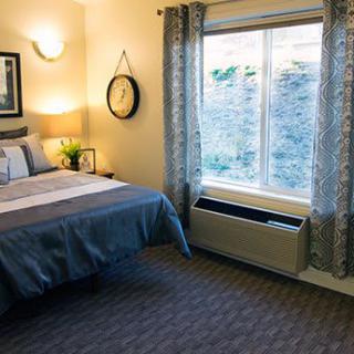 A cozy bedroom with a neatly made bed, wall-mounted clock, and large window letting in natural light.