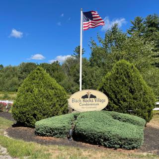 Entrance sign with an American flag surrounded by landscaped bushes and trees on a sunny day.