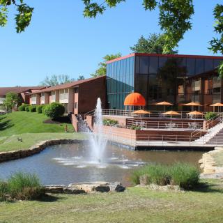 A building with outdoor seating overlooks a pond adorned with a fountain under a clear blue sky.