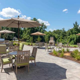 A spacious outdoor patio with tables, chairs, and umbrellas set against a lush garden backdrop on a sunny day.
