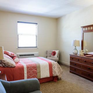 A cozy bedroom with a neatly made bed, a dresser with a mirror, and a chair by the window.