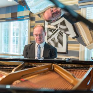 A man in a suit playing the piano indoors with his reflection visible.
