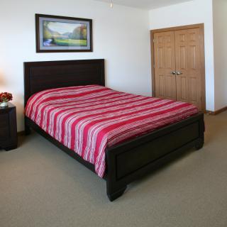 A neatly arranged bedroom featuring a bed with red striped bedding, a nightstand with a lamp, and a landscape painting on the wall.