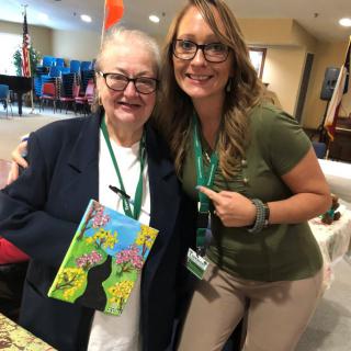 Two women smiling, one holding a colorful book indoors.