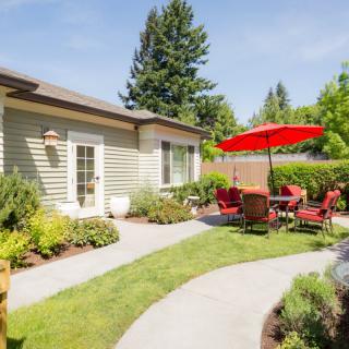 A well-manicured backyard featuring a patio set with a red umbrella, surrounded by lush greenery and a pathway.