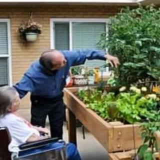 A person in a wheelchair observing another person tending to plants in a raised garden bed.