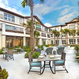 A sunny outdoor patio area with tables and chairs set in front of a modern residential building, surrounded by palm trees.