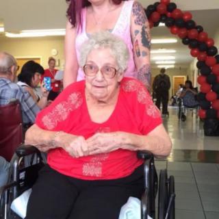 An elderly woman in a wheelchair is being assisted by a caregiver at an indoor event decorated with balloons.
