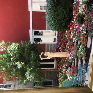 A peaceful courtyard with a statue surrounded by vibrant flowers and greenery against a red wall.