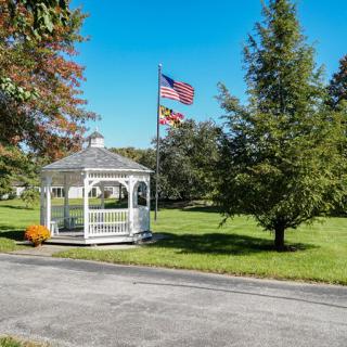 A scenic view of a white gazebo in a garden, surrounded by trees and flying flags under a clear blue sky.