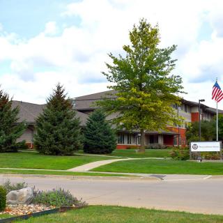 A two-story office building surrounded by trees and a well-kept lawn, featuring an American flag and a sign at the entrance.