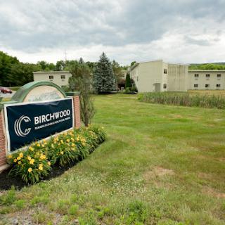 A view of the Birchwood facility surrounded by lush greenery and blooming flowers under a cloudy sky.