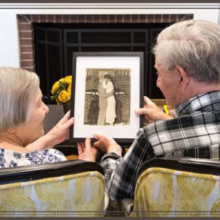 An elderly couple sits together in their living room, cherishing a framed photograph from the past.
