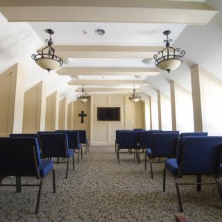 A small chapel with rows of chairs, a cross on the wall, and elegant lighting fixtures.