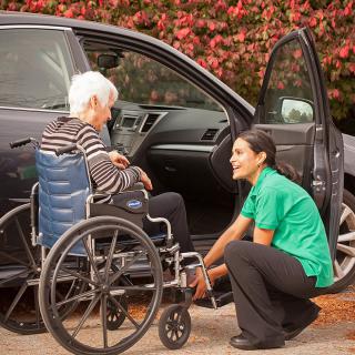 A caregiver assists an elderly person in a wheelchair next to a car.