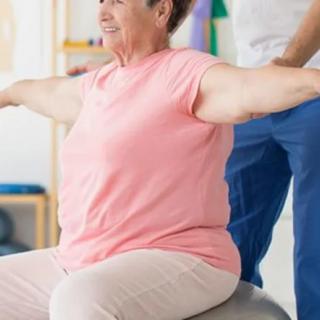 An elderly woman performing a balance exercise with the assistance of a trainer.