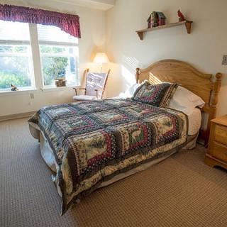 A cozy bedroom with a wooden bed frame and a colorful quilt, featuring natural light from the windows.