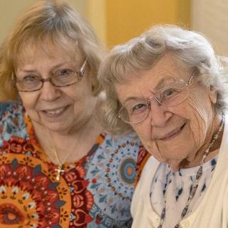 Two elderly women smiling and sitting together indoors.