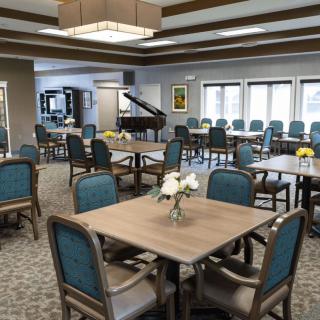 A spacious community dining room with neatly arranged tables and chairs, featuring a grand piano in the corner.