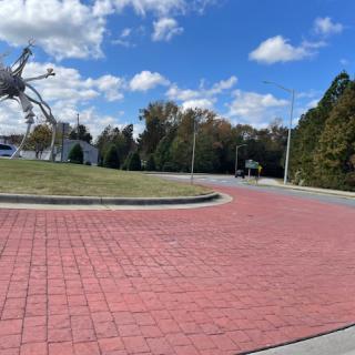 A scenic outdoor area featuring a large sculpture, surrounded by trees and a paved pathway under a partly cloudy sky.