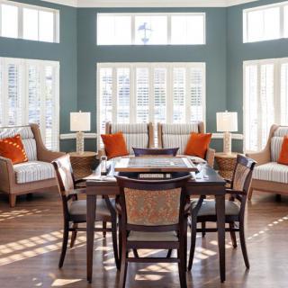 A modern dining room with large windows, featuring wooden furniture and orange accent pillows.