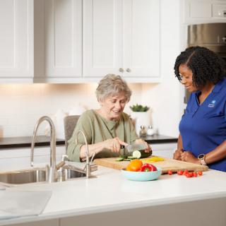 An elderly individual prepares food with the assistance of a caregiver in a bright kitchen.