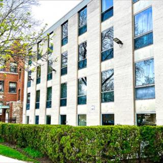A modern residential apartment building with large windows and a neatly trimmed hedge lining the sidewalk.