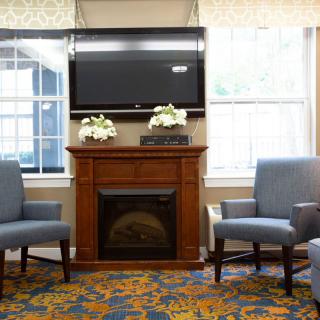 A cozy living room with a fireplace and a TV above, flanked by cushioned chairs and decorated with floral arrangements.