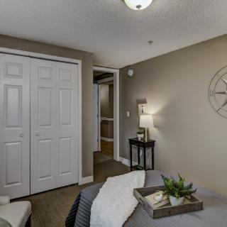 A cozy bedroom with neutral tones featuring a bed, a bedside lamp on a small table, closet doors, and decorative wall art.