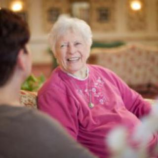 An elderly woman wearing a pink sweater is smiling while having a conversation.
