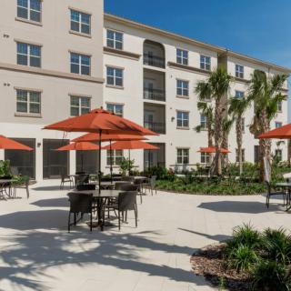 A sunny outdoor patio area with tables, chairs, and orange umbrellas in front of an apartment building surrounded by palm trees.