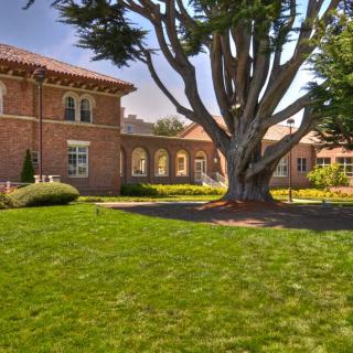 A scenic view of a brick building surrounded by lush greenery and a large tree on a sunny day.
