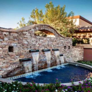 A scenic outdoor fountain with a stone wall and water cascading into a pool, surrounded by lush garden landscaping.