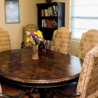 A cozy dining room with a round wooden table and upholstered chairs, featuring a vase of colorful flowers and a bookshelf in the background.