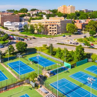 Aerial view of a series of blue tennis courts surrounded by urban buildings and parking lots.