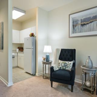 A cozy living area with a view into a small kitchen, featuring a comfortable black chair and side tables with a lamp and decor.