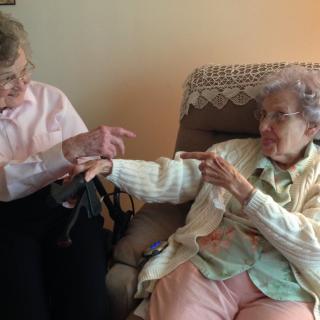 Two elderly women sitting together and pointing at each other while conversing.